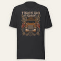 Trucking Cas Tee in black heather, front view laid flat showing an orange vintage big rig under a TRUCKING Apparel script headline with ornate scrollwork and American flag accents.